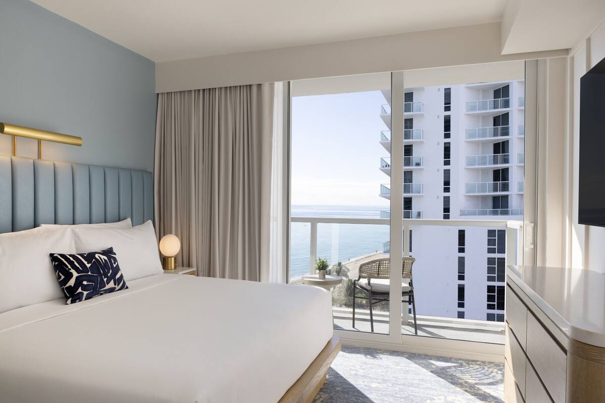 Habitación de hotel con cama, mesita de noche, televisor, balcón y vista al mar y un edificio.