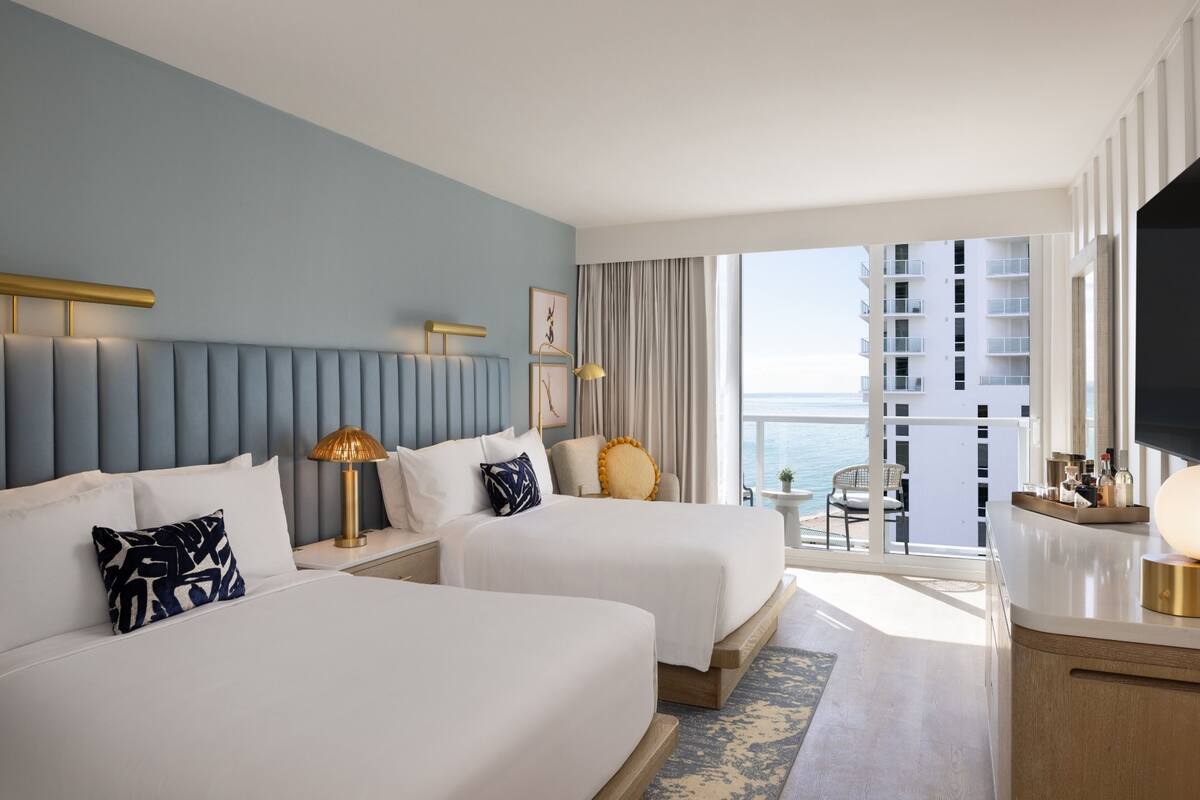 Habitación de hotel con dos camas, televisión, balcón y vistas al mar, además de un área de comedor.