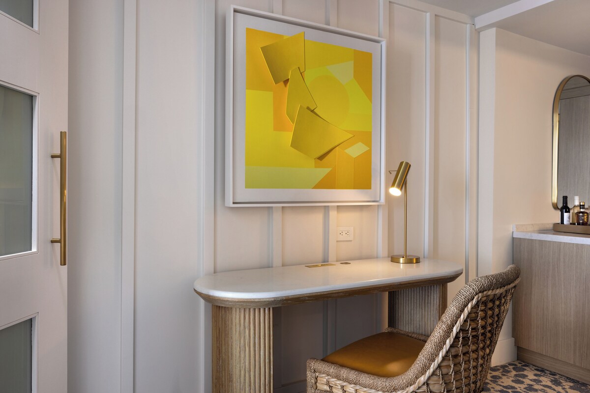 A room with a white console table, a wicker chair, a framed abstract painting, and a mirror above the console table.