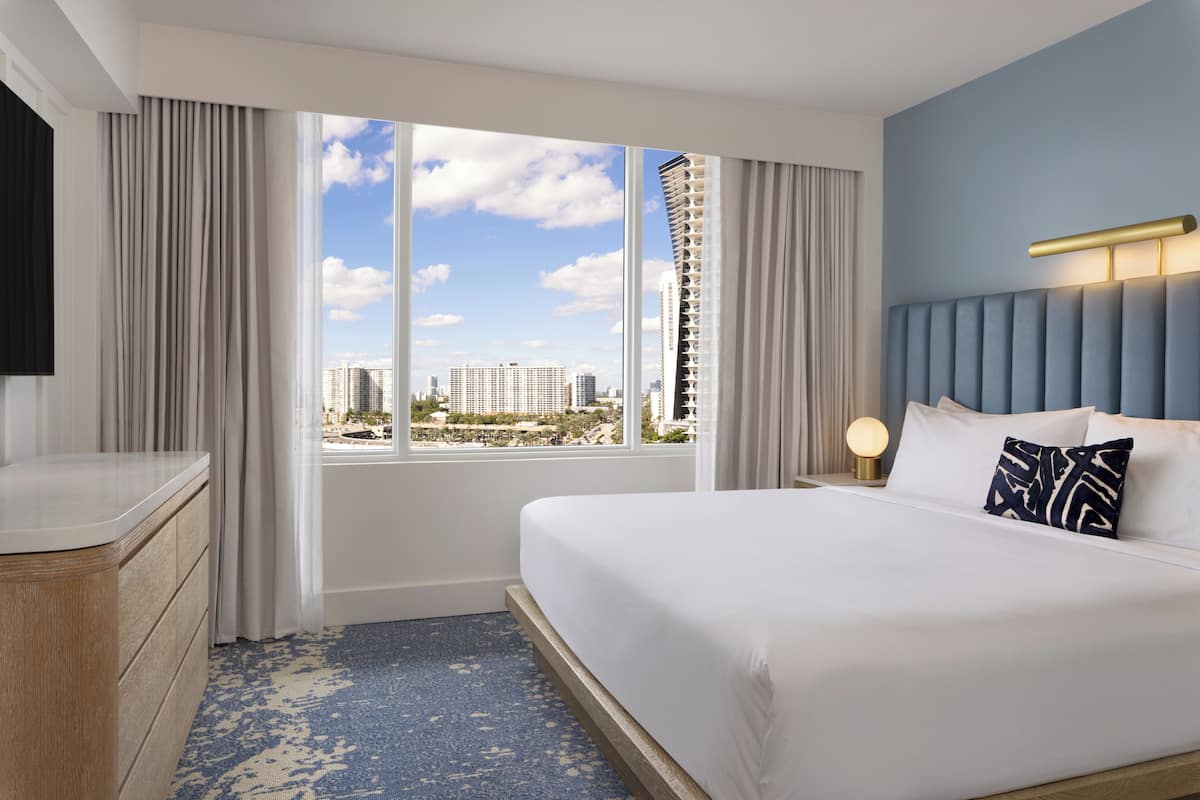 Habitación de hotel con una cama grande, una mesita de noche con lámpara, cómoda y vistas a la ciudad a través de amplios ventanales.