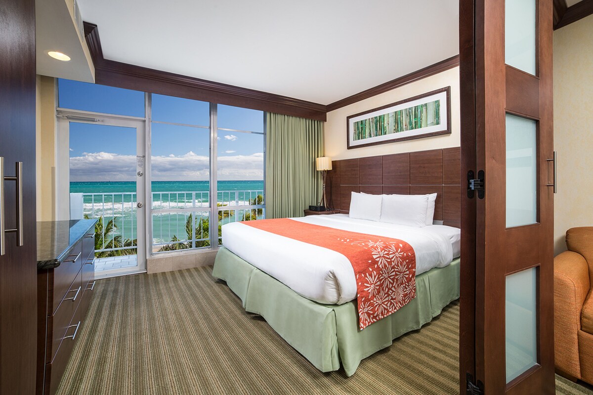 A hotel room with a large bed, a view of the ocean, a balcony, and a desk.