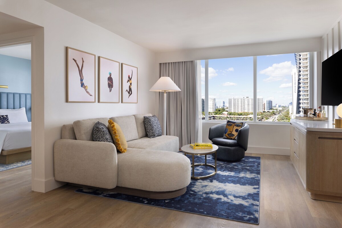 A modern hotel room with a sofa, armchair, small round table, and a large window with a city view.