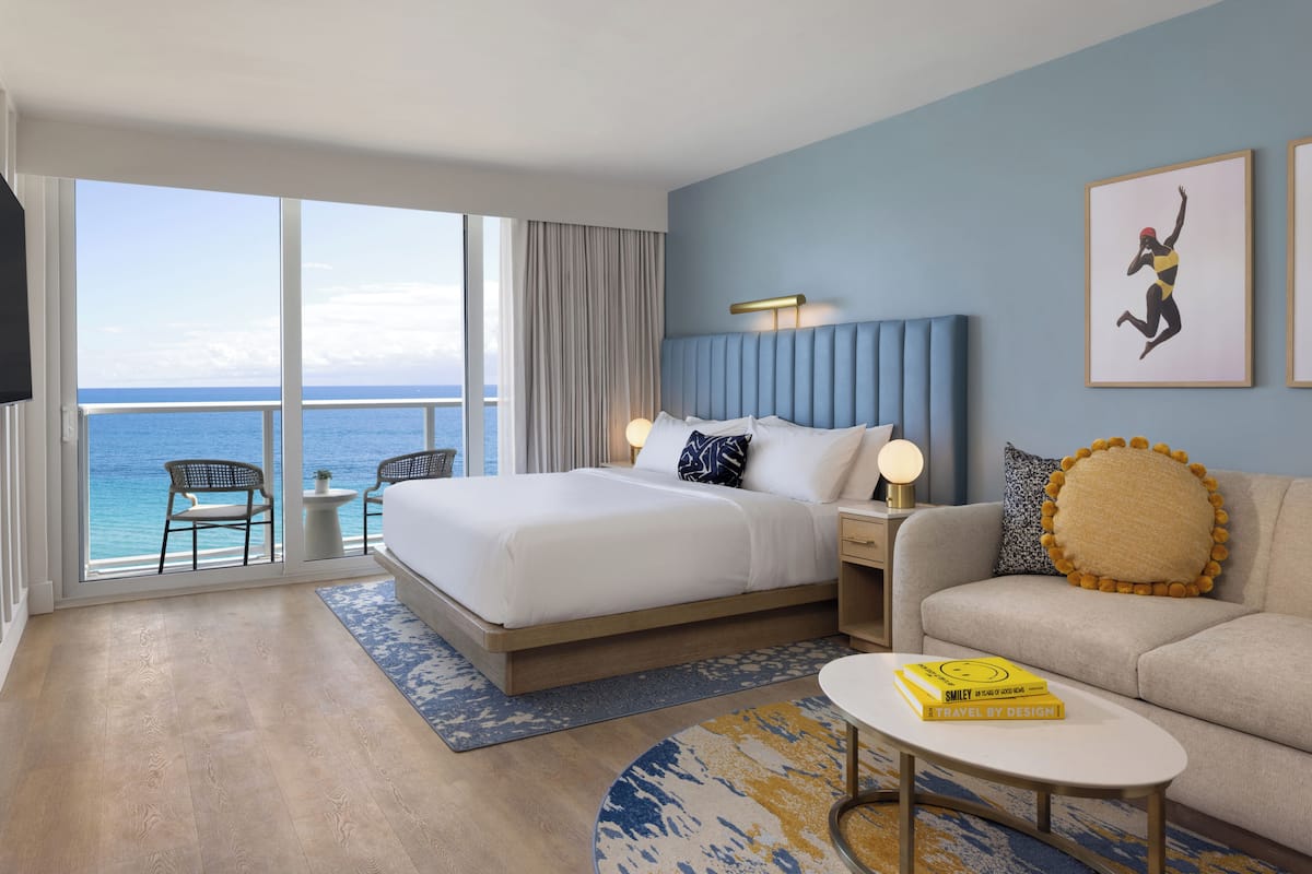 Habitación de hotel con cama, sofá, mesa de centro, televisor, balcón con vista al mar y una obra de arte enmarcada en la pared.