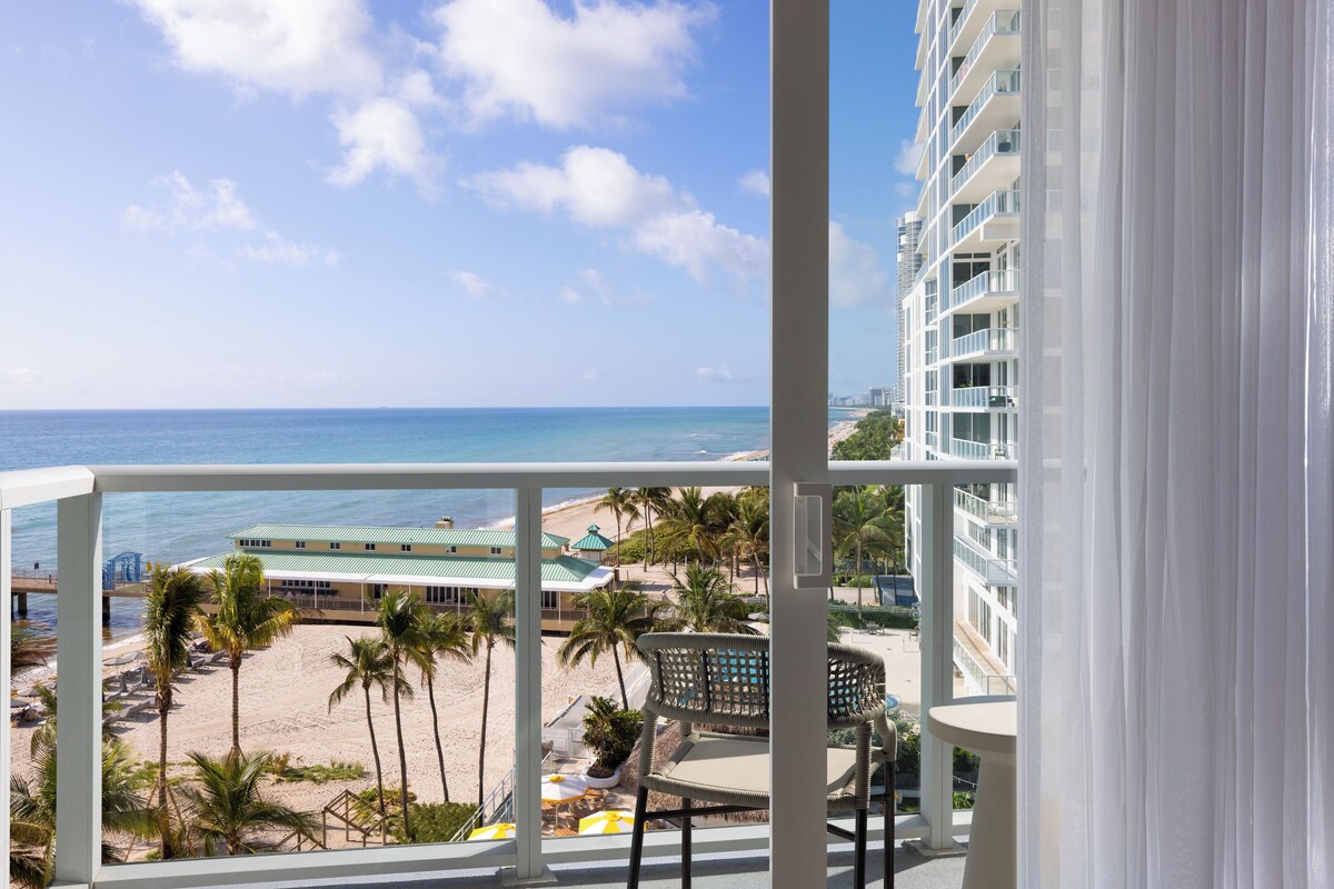 A balcony with a view of a beach, palm trees, and a building with a green roof.