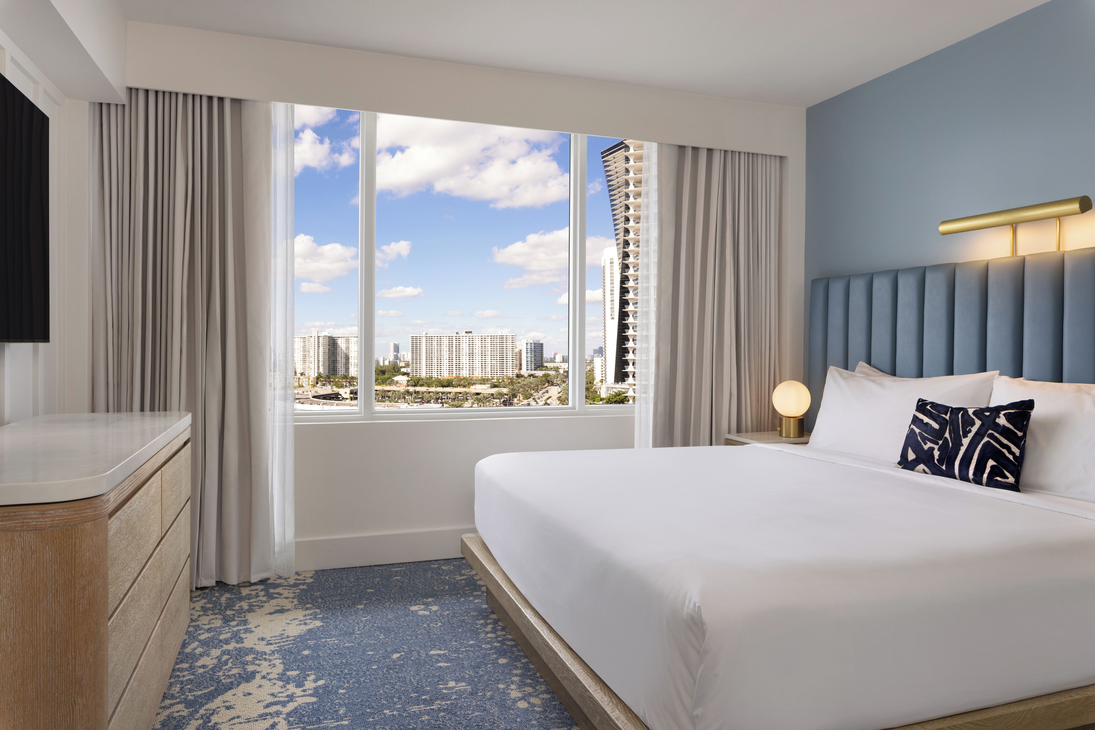 Habitación de hotel con una cama grande, una mesita de noche con lámpara, cómoda y vistas a la ciudad a través de amplios ventanales.