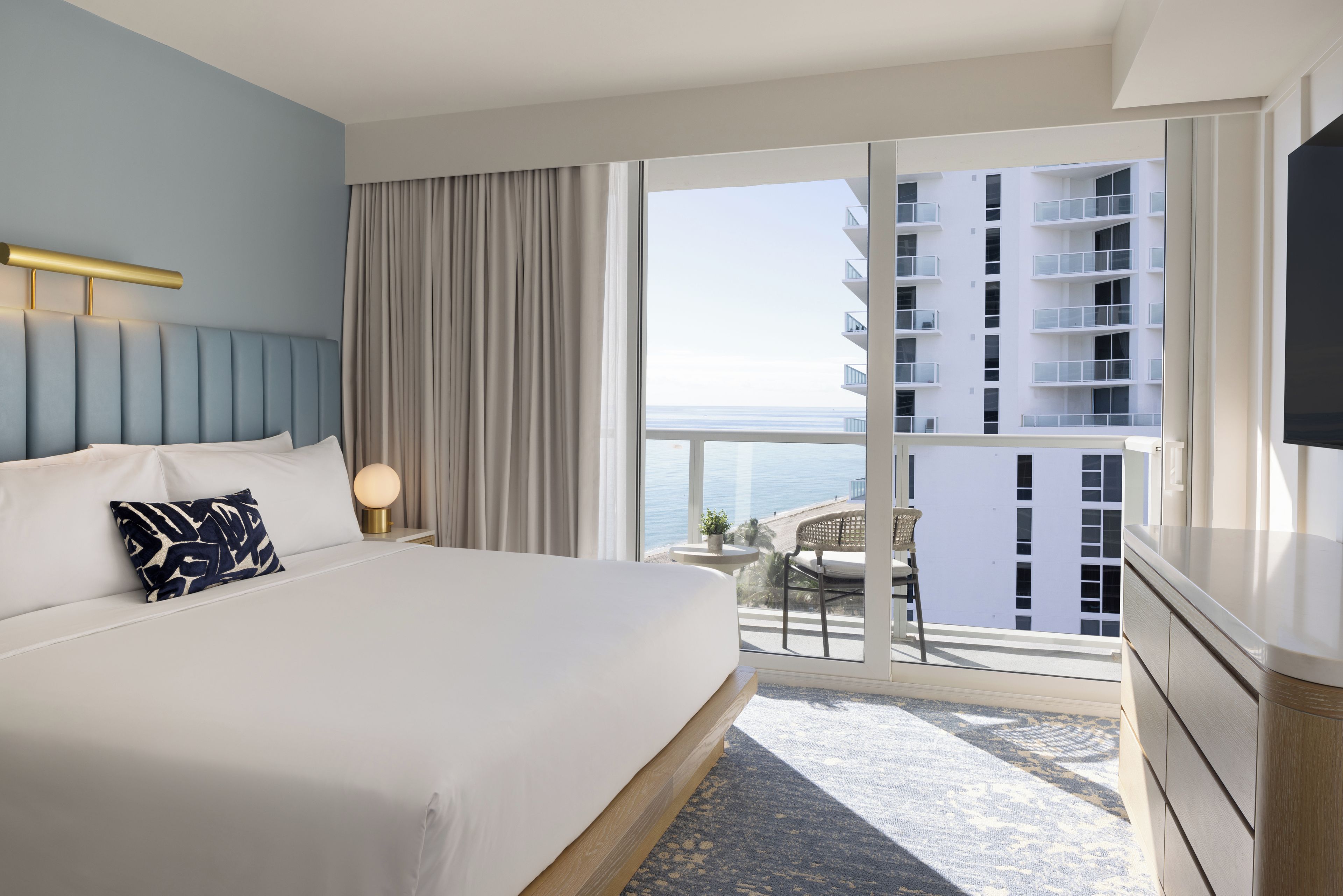 Ein Hotelzimmer mit Bett, Nachttisch, Lampe, Fernseher und einem Balkon mit Blick auf das Meer und Gebäude.
