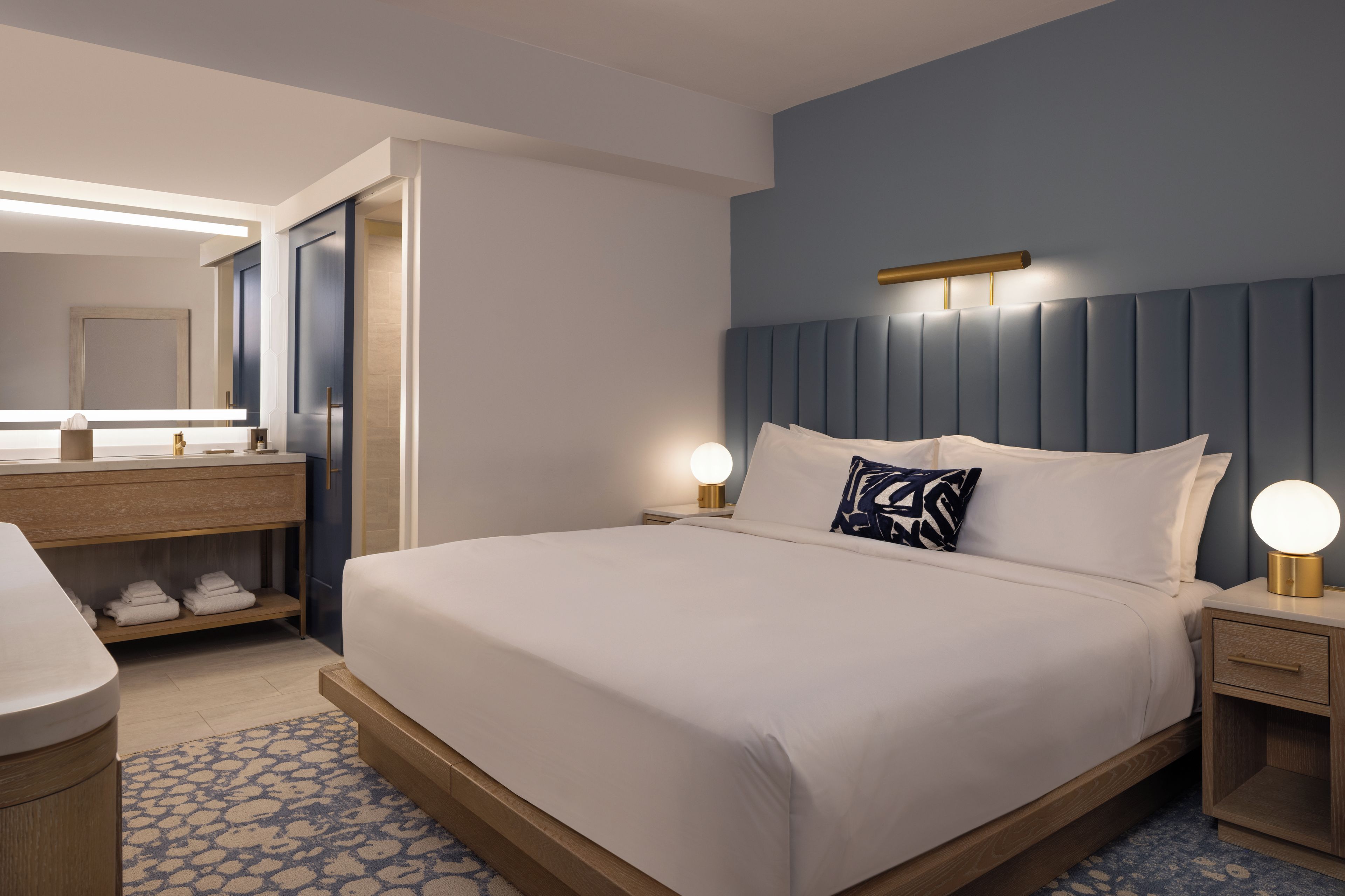 A hotel room with a large bed, bedside lamps, a wooden nightstand, a built-in shelf with towels, and a mirror above the vanity.