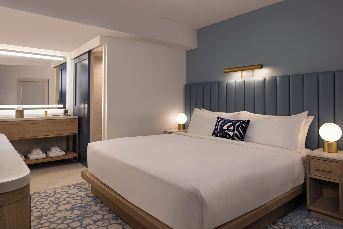 A hotel room with a large bed, bedside lamps, a wooden nightstand, a built-in shelf with towels, and a mirror above the vanity.