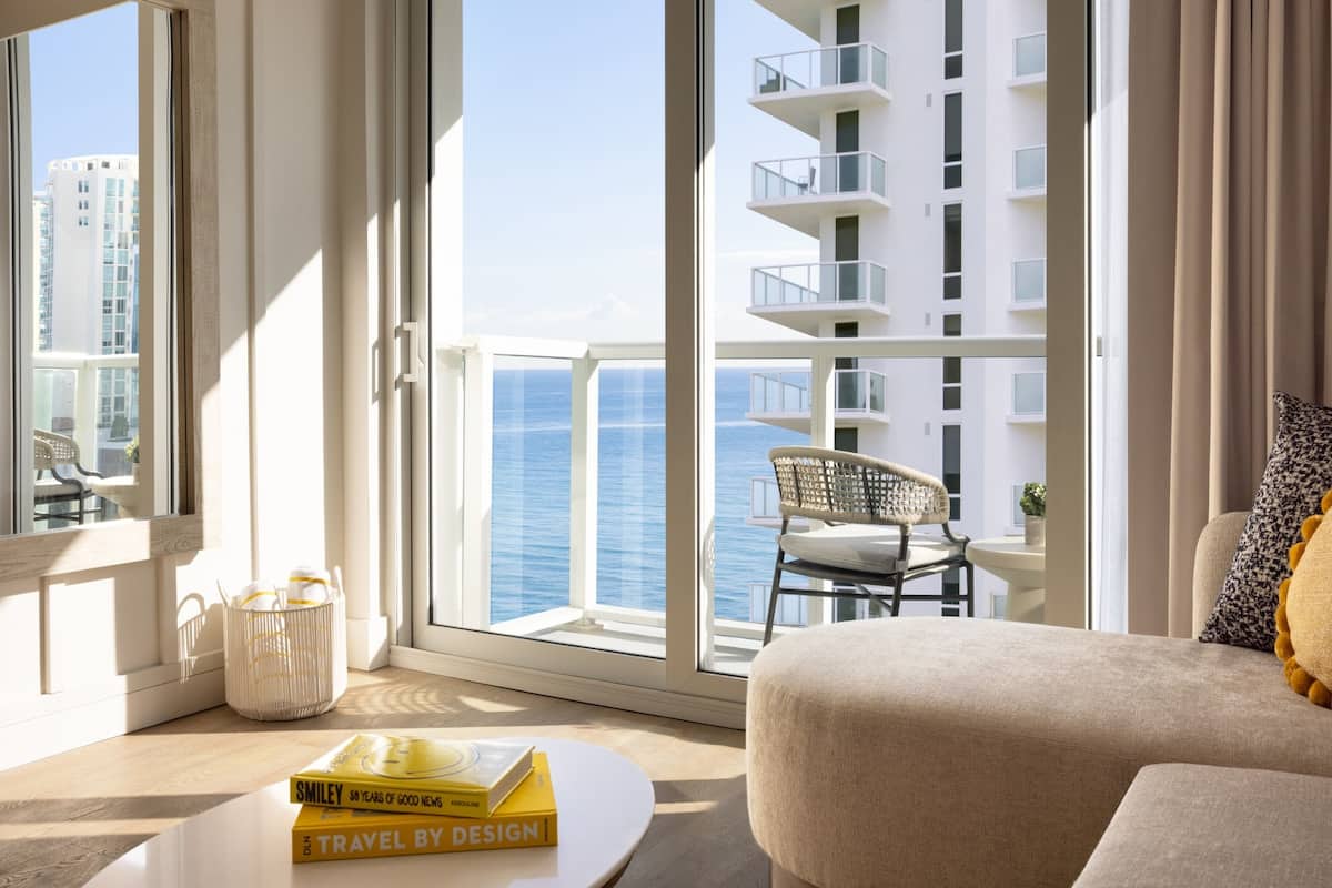 Una sala de estar moderna con vistas al mar, un balcón con una silla y una pila de libros sobre una mesa de centro.