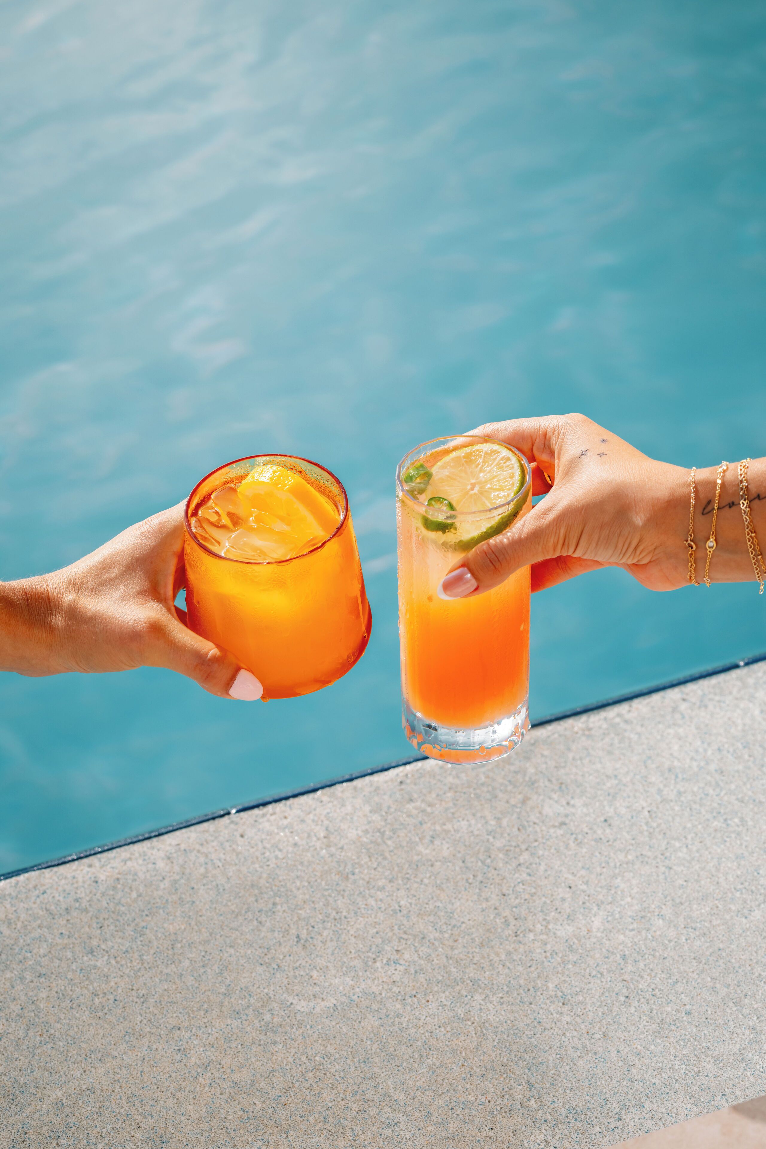 Two hands holding glasses of orange juice with lime slices by a pool.