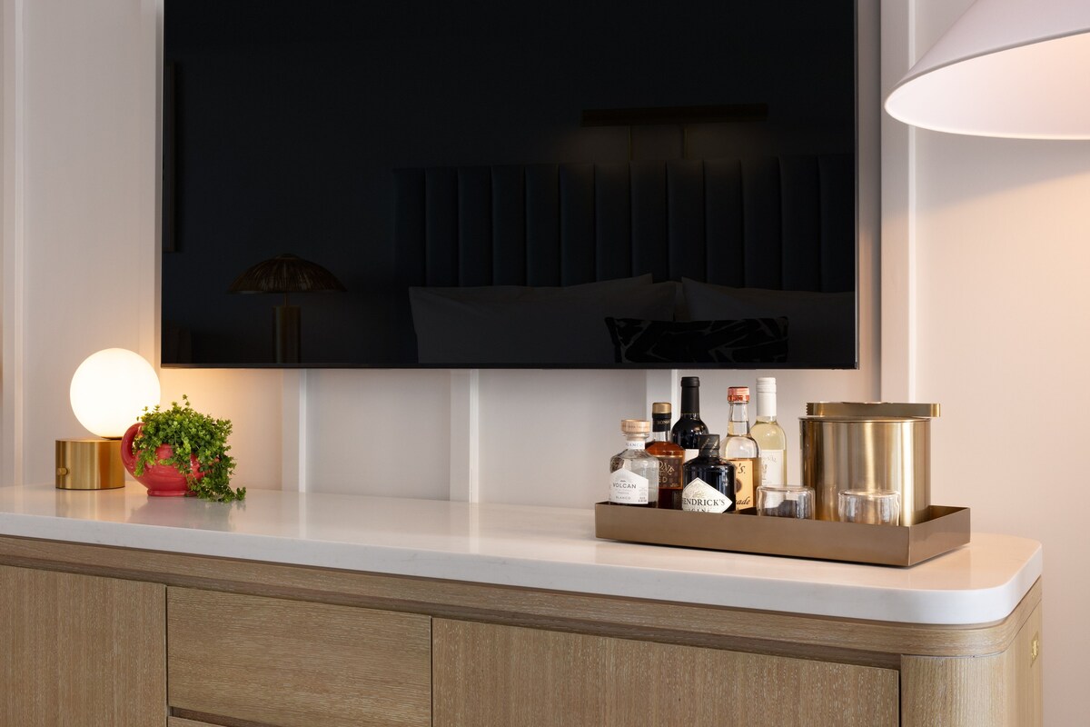 A modern hotel room with a large flat-screen TV mounted on the wall, a bedside lamp, a small potted plant, and a tray of toiletries.