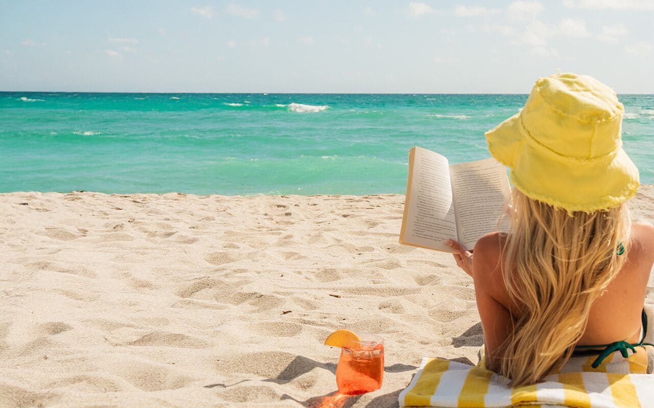 A person reading a book on a beach with a drink and a towel.