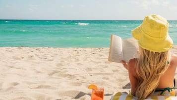 Una persona leyendo un libro en la playa, con una bebida y una toalla.