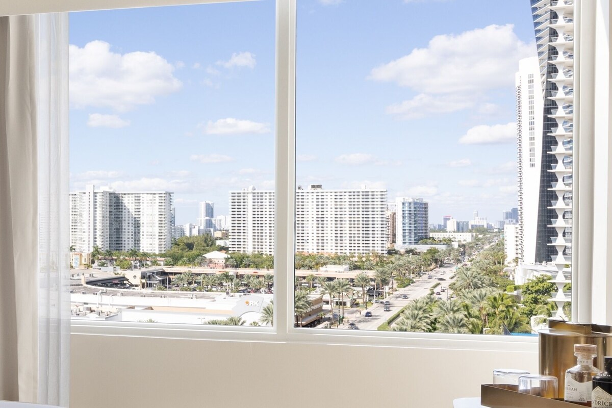 A hotel room with a large window, a bed, a nightstand, and a view of a cityscape with tall buildings.