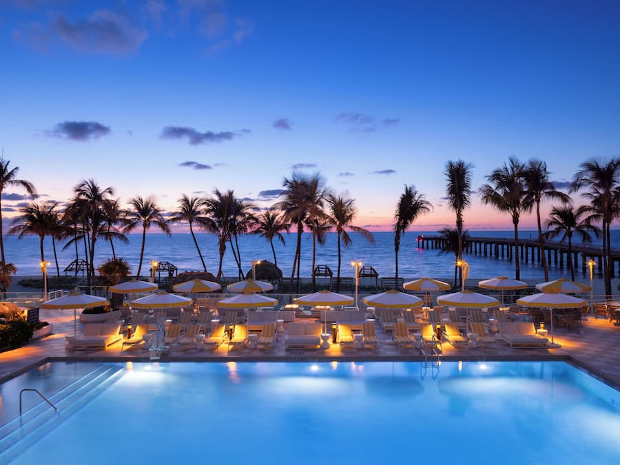 A poolside area with lounge chairs and umbrellas, palm trees, and a view of the ocean at dusk.