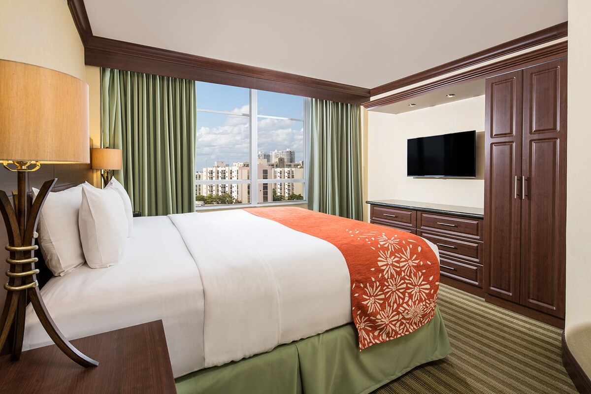 A hotel room with a bed, a TV, a desk, and a window with curtains.