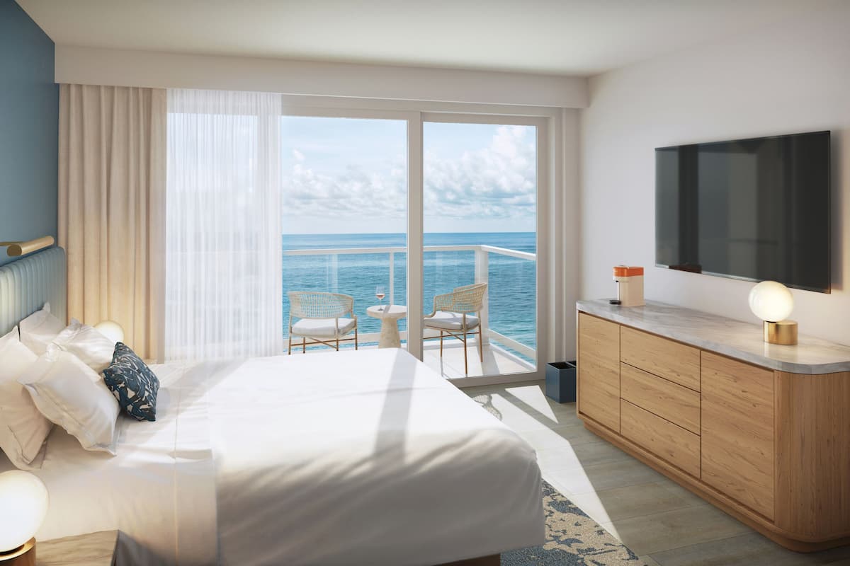 Habitación de hotel con cama, televisión, balcón con vista al mar y una pequeña mesa con sillas.