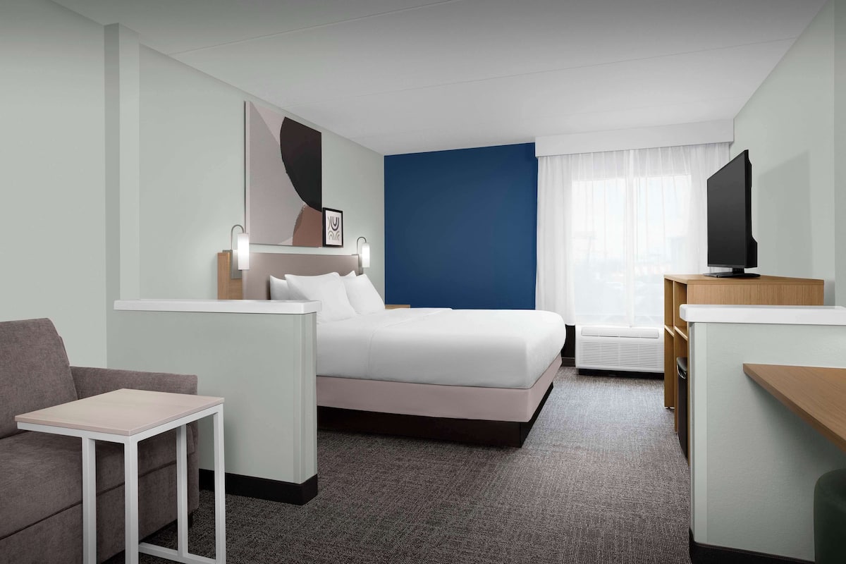 Une chambre d’hôtel avec un lit, une petite table, un canapé, une télévision et une fenêtre avec des rideaux.