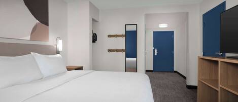 Habitación de hotel con una cama grande, un escritorio con televisor, un espejo y una puerta.