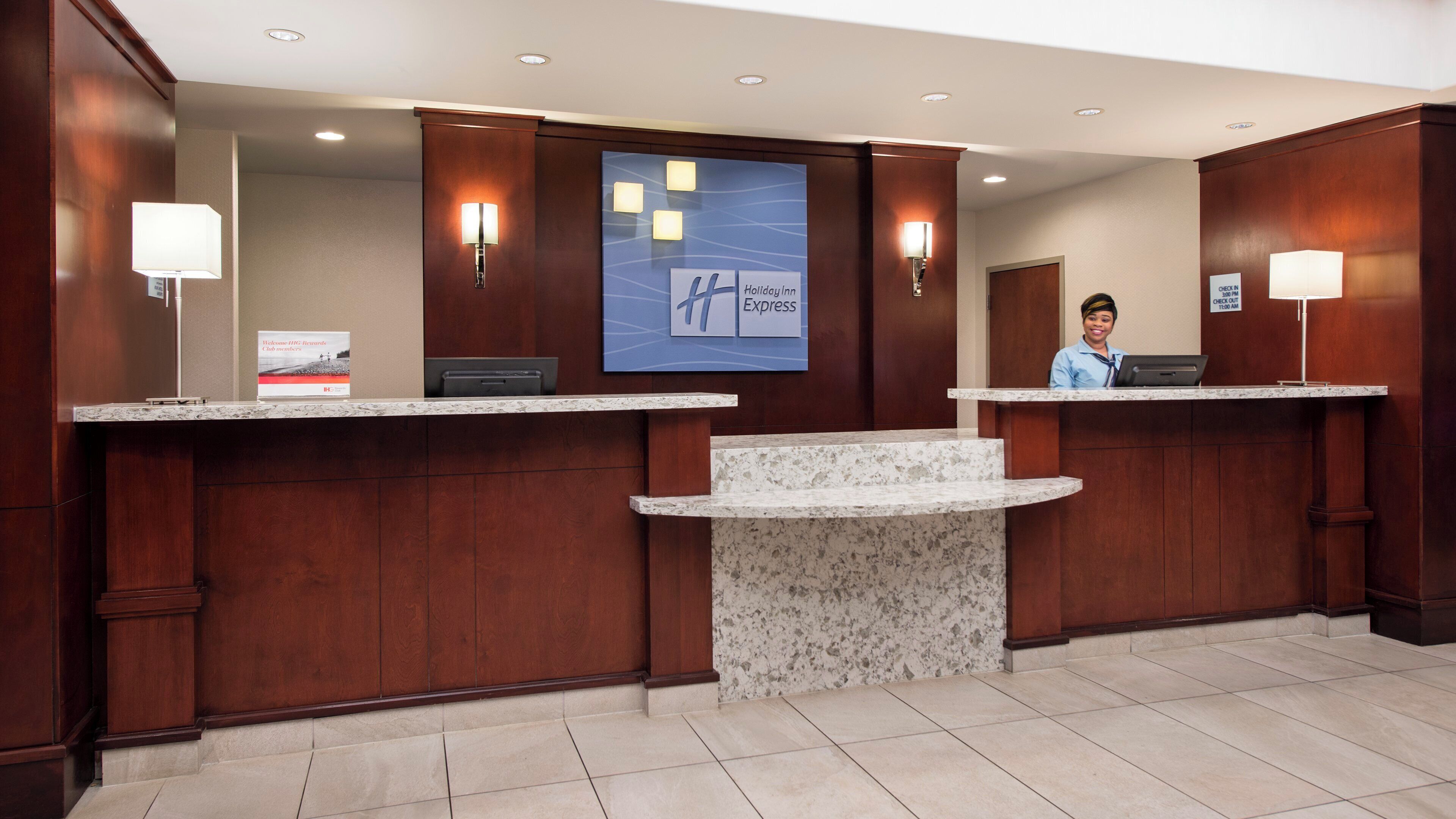 A hotel reception area with two counters, a large screen displaying the hotel name, and a sign indicating check-in times.
