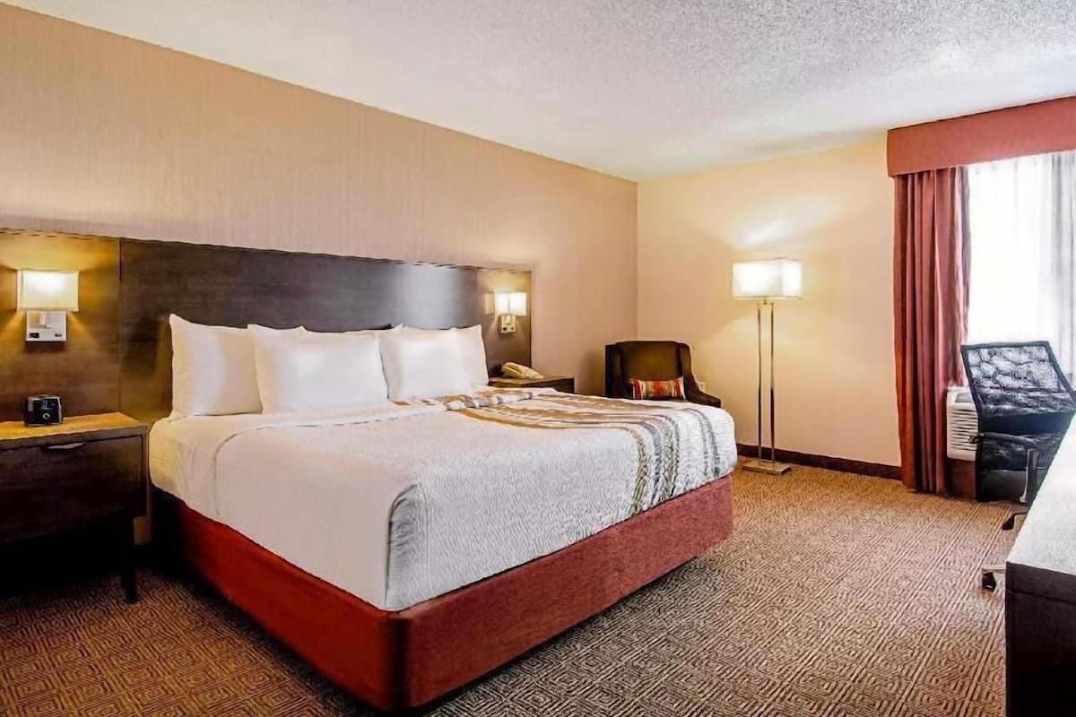 Una habitación de hotel con una cama grande, lámparas de noche, una silla, un escritorio y un ventanal con cortinas.