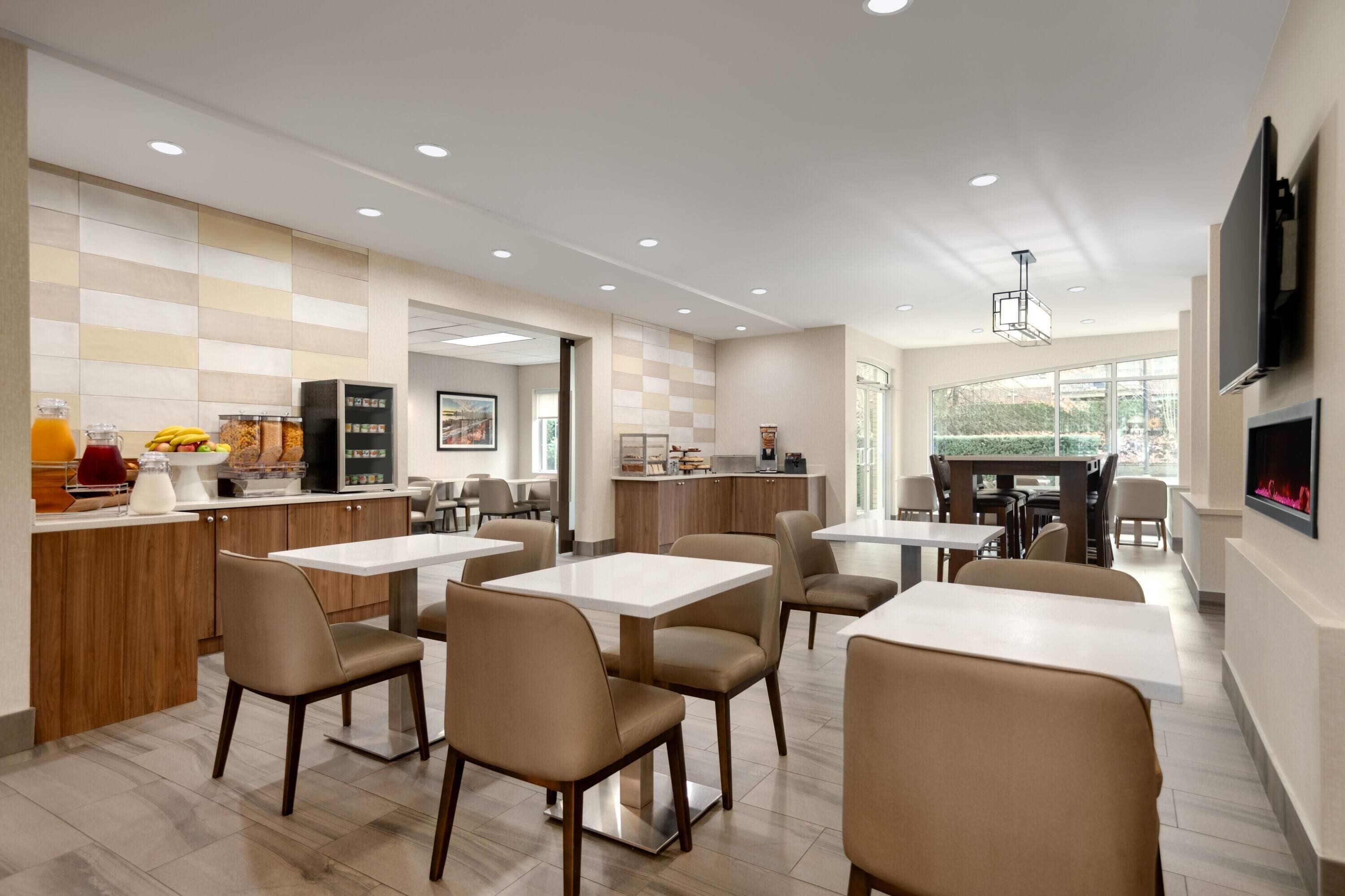 A modern dining area with multiple tables and chairs, a kitchen area with a refrigerator and counter, and a large window providing natural light.