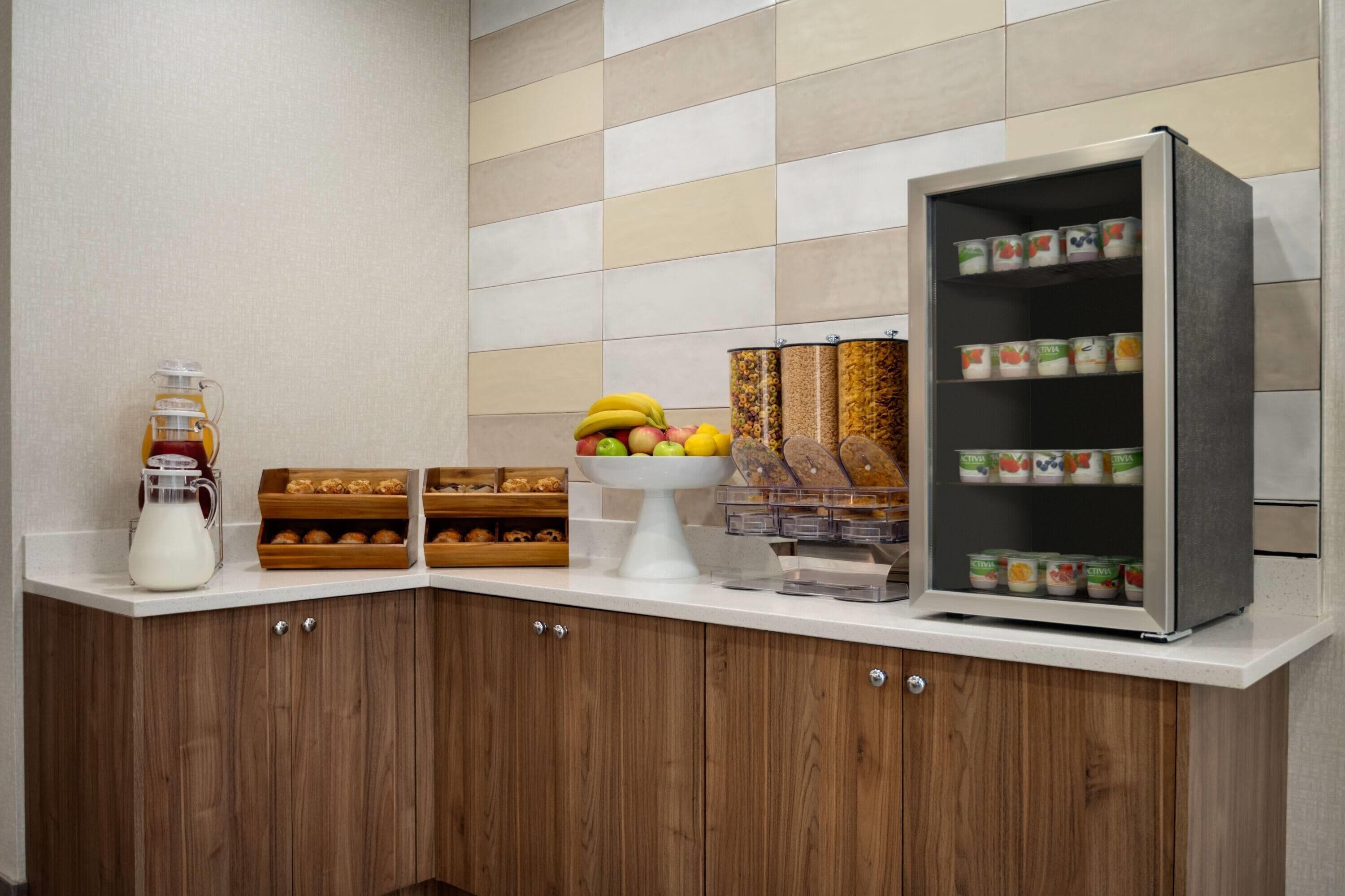 A breakfast bar with a milk pitcher, fruit bowl, bread, and a refrigerated display.