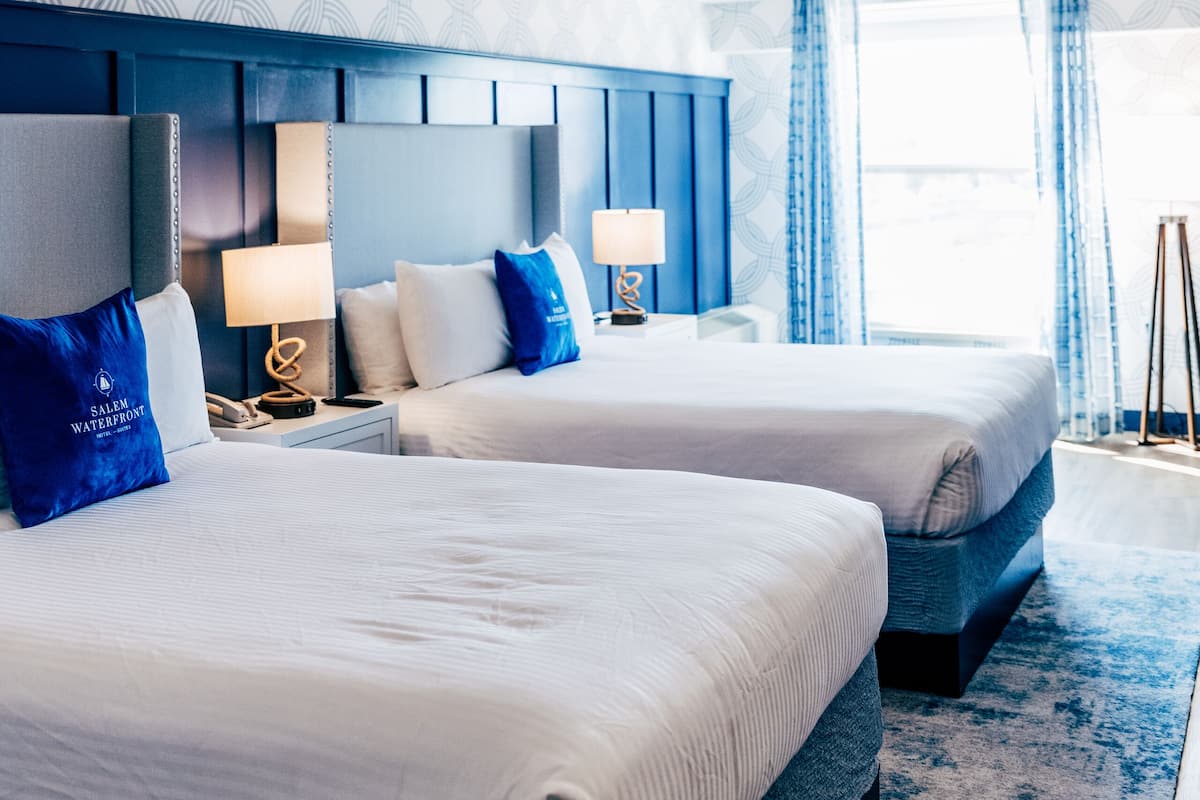 Habitación de hotel con dos camas, cabecera azul, pared con diseño y una ventana grande con cortinas.