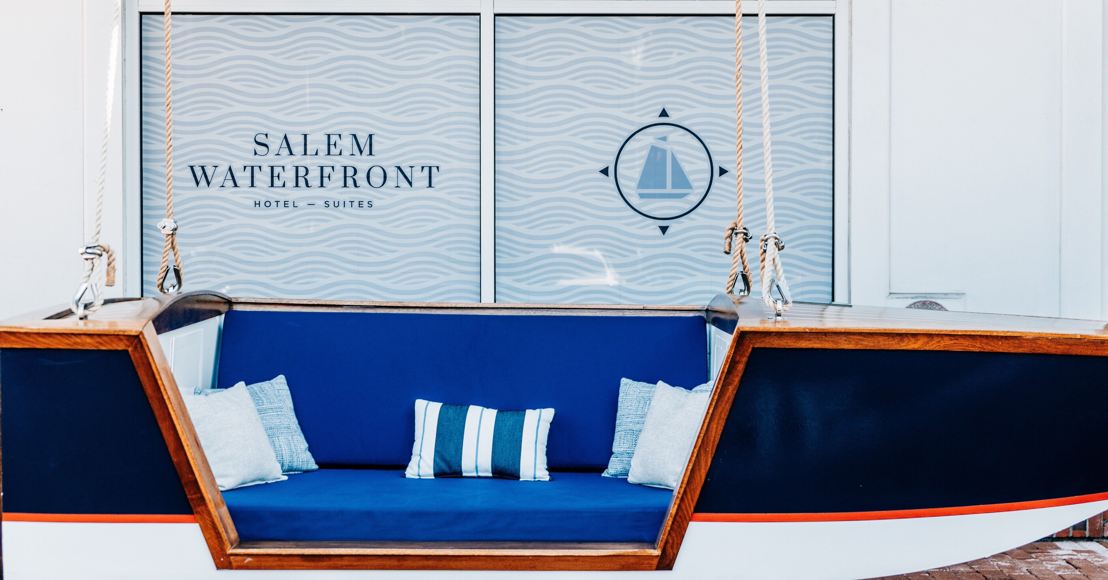 Ein blaugrauer Polsterschaukelsitz mit weißen und blauen Kissen vor einer Wand mit einem Wellenmuster und dem Schriftzug 'SALEM WATERFRONT HOTEL - SUITES'.