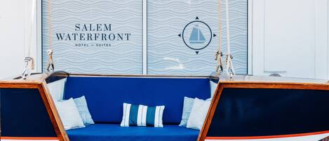 Ein blaugrauer Polsterschaukelsitz mit weißen und blauen Kissen vor einer Wand mit einem Wellenmuster und dem Schriftzug 'SALEM WATERFRONT HOTEL - SUITES'.