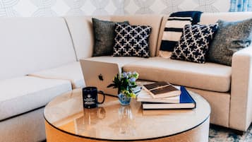 A modern living room with a beige sofa, patterned cushions, a glass coffee table, a laptop, a blue mug, a small plant, and a book.