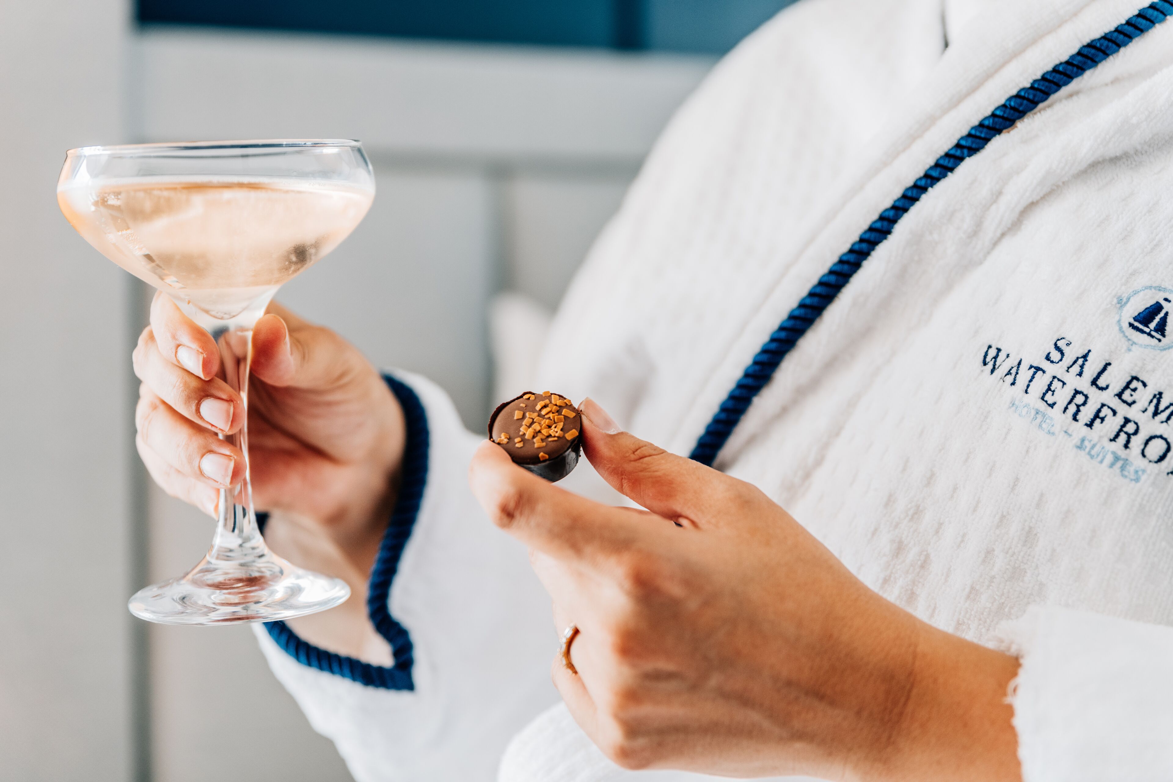 Una persona sosteniendo una copa de martini y un dulce cubierto de chocolate. La persona lleva una bata con la inscripción "Salem Waterfront Hotel".