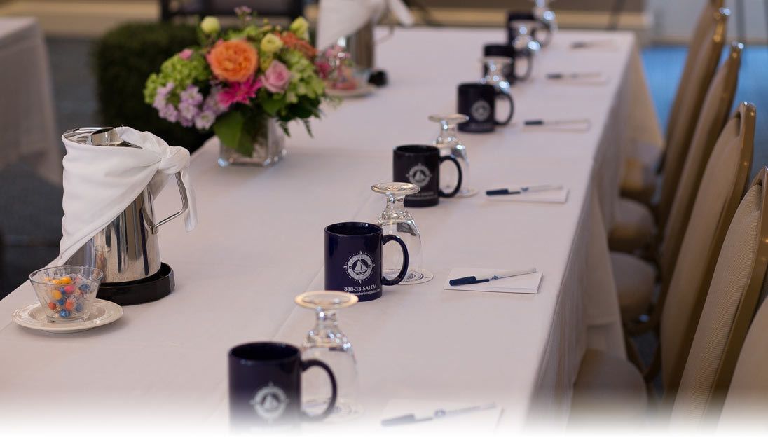 A long table with a white tablecloth, black mugs, and a silver teapot.