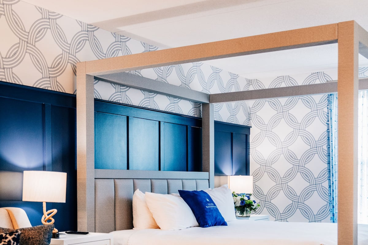 Una habitación de hotel moderna con una cama grande, una mesita de noche con lámpara, una mesa de centro redonda y vistas a la ciudad desde la ventana.
