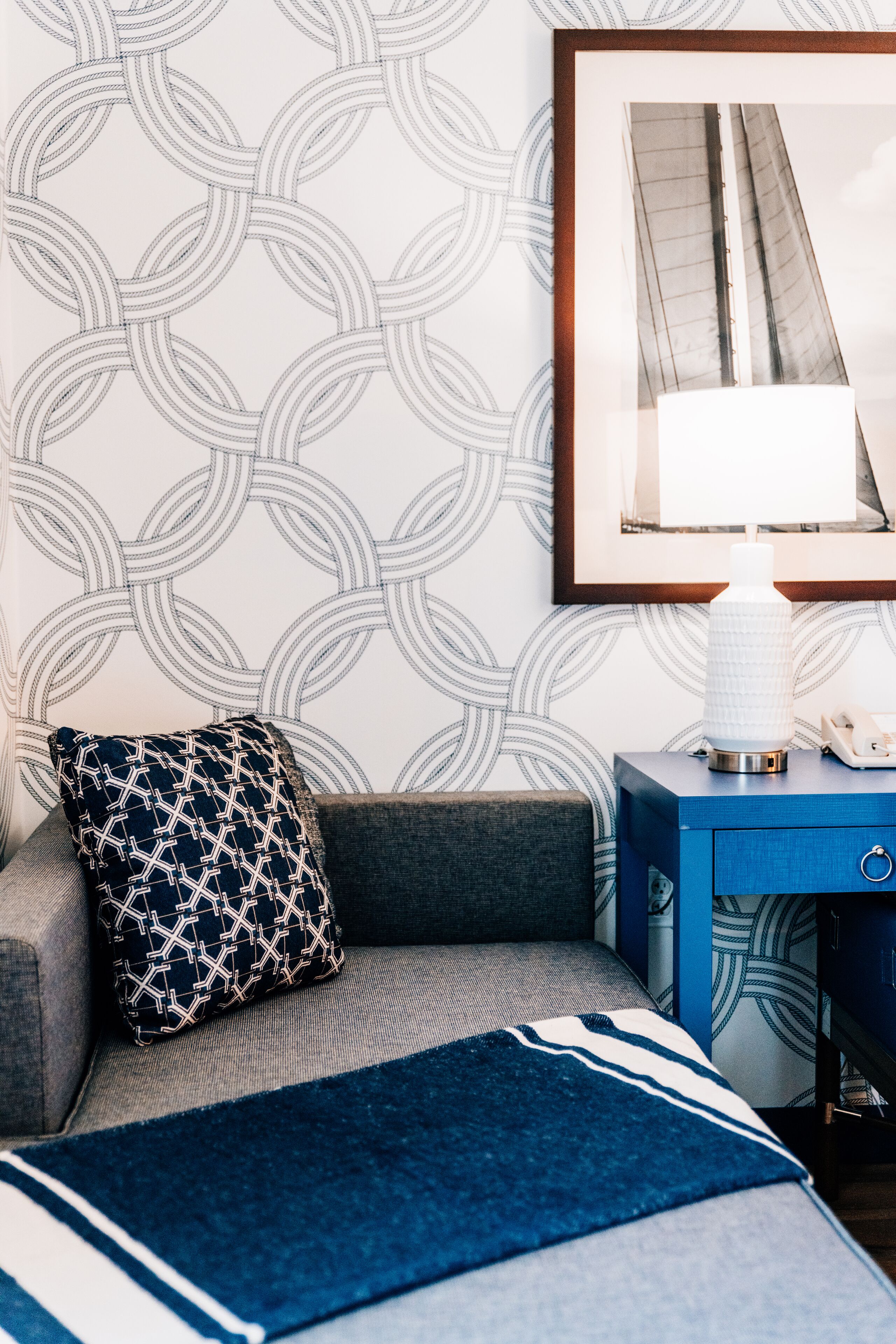 A neatly arranged bedroom with a patterned wallpaper, a bedside lamp, a patterned pillow, and a blue bedside table.