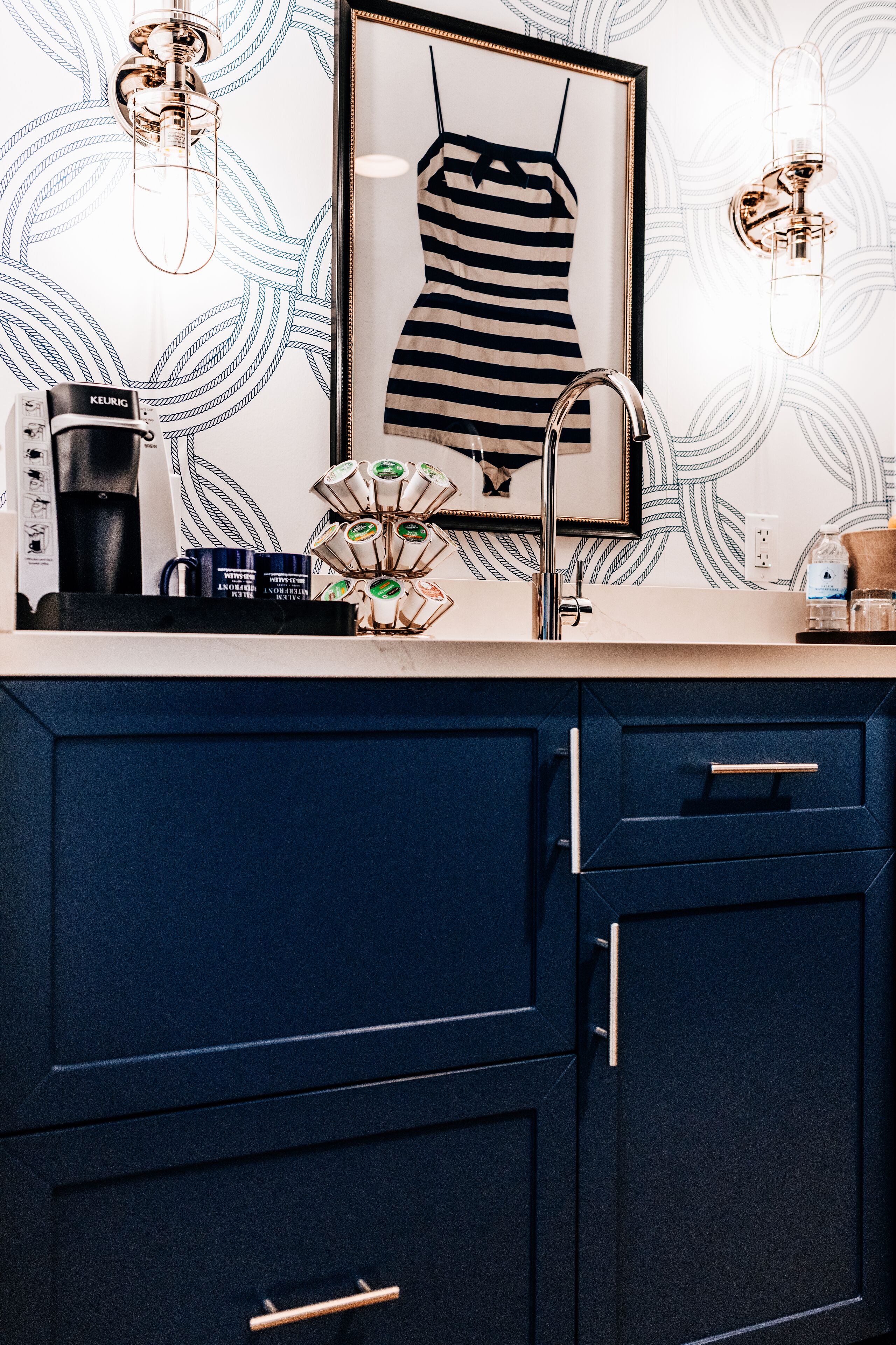 A kitchen with dark blue cabinets, a coffee maker, a framed picture on the wall, and a patterned wallpaper.