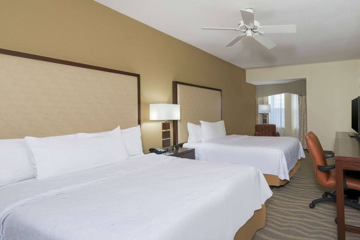 Ein Hotelzimmer mit zwei Betten, einem Schreibtisch, einem Sessel, einem Fernseher und einem Deckenventilator.