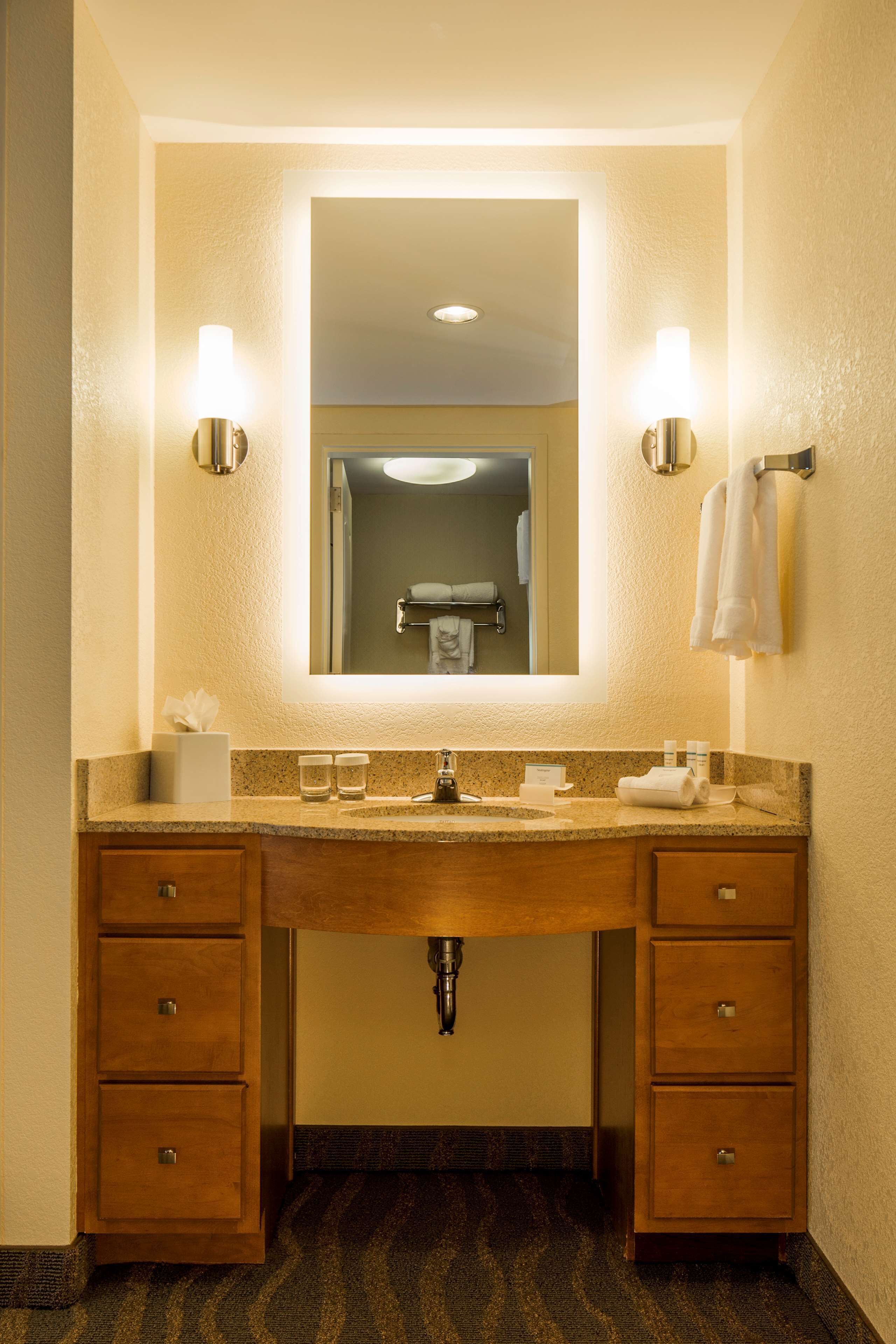 A hotel bathroom with a wooden vanity, two wall-mounted lights, a mirror, and a towel rack.