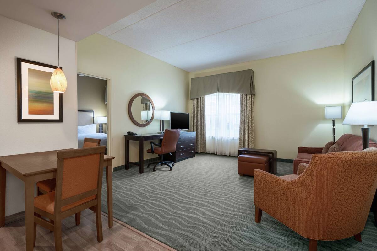 Ein Hotelzimmer mit Schreibtisch aus Holz, einem Sessel, einer Couch, einem Fernseher, einem Spiegel und einem gerahmten Bild an der Wand.