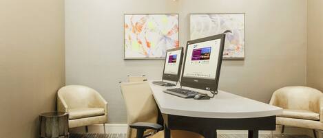 Un salón de conferencias con dos monitores de computadora, una mesa blanca, dos sillas color beige, un cesto de basura pequeño y una obra de arte abstracta en la pared.