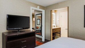 A hotel room with a flat-screen TV on a dark wooden dresser, a bed with white bedding, a door leading to a bathroom, and a small mirror on the wall. - Homewood Suites Melville (Melville)