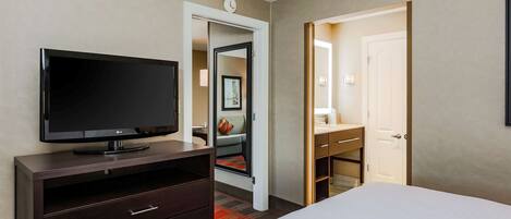 Habitación de hotel con televisor de pantalla plana sobre un aparador de madera oscura, cama con ropa de cama blanca, puerta que da a un baño y un espejo pequeño en la pared.