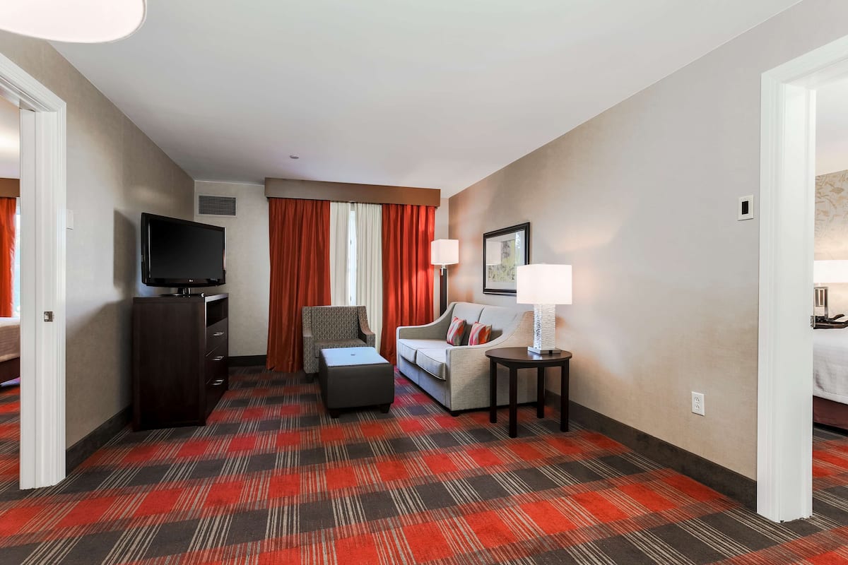 A hotel room with a flat-screen TV, a sofa, a chair, a small table with a lamp, and a patterned carpet.