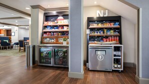 A self-service beverage station with refrigerated drinks and snacks, located in a modern hotel lobby with wooden flooring and seating areas. - Hampton Inn Princeton (Princeton)