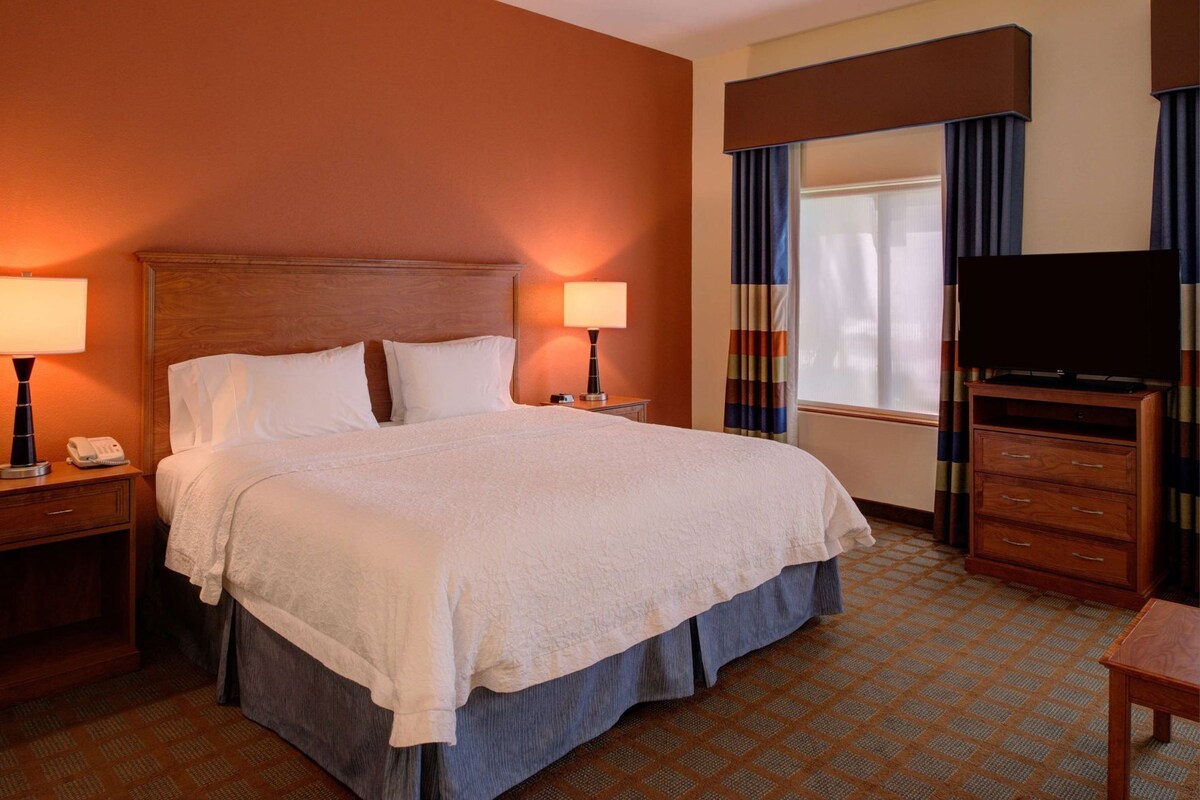 A hotel room with a large bed, two bedside lamps, a TV, a wooden dresser, and a window with curtains.