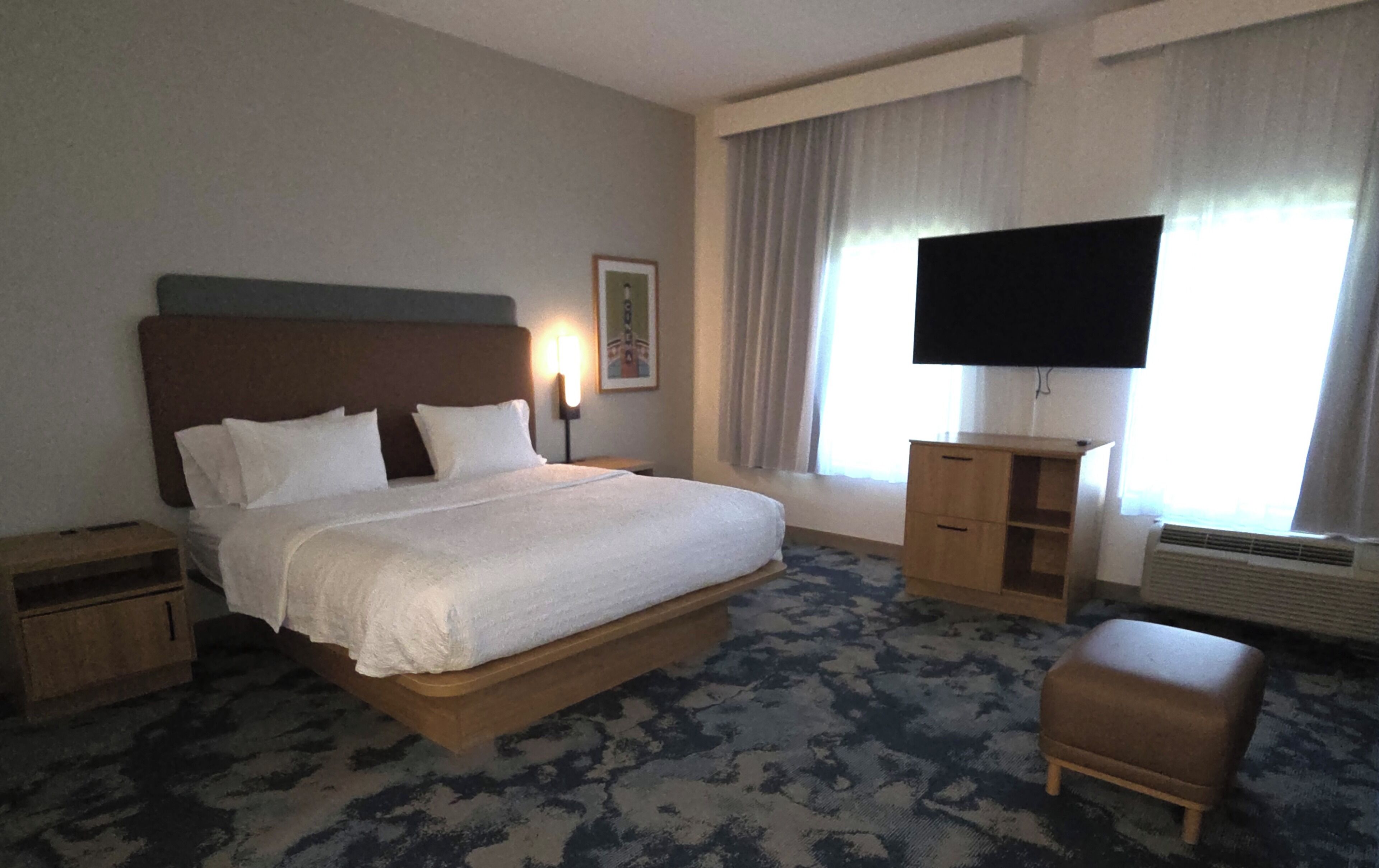 A hotel room with a large bed, a flat-screen TV, a wooden nightstand, a wooden dresser, and a bench.