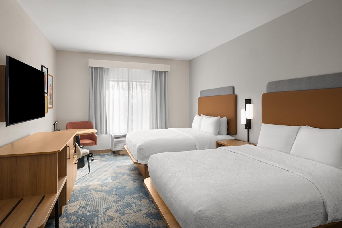 A hotel room with two beds, a desk, a chair, a TV, and a window with curtains.