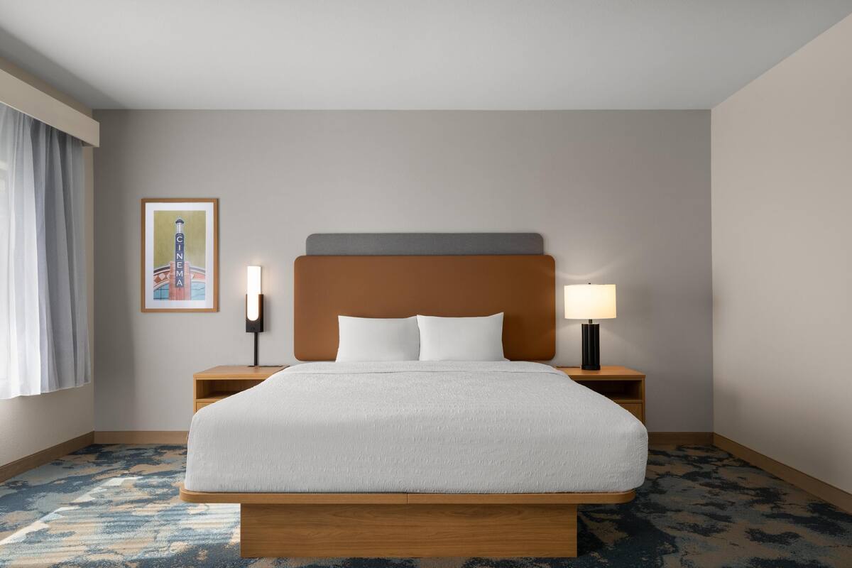 A hotel room with a large bed, a nightstand with a lamp, a framed picture on the wall, and a window with curtains.