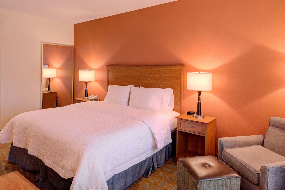 A hotel room with a large bed, two bedside tables with lamps, a wooden headboard, a small armchair, and an ottoman.