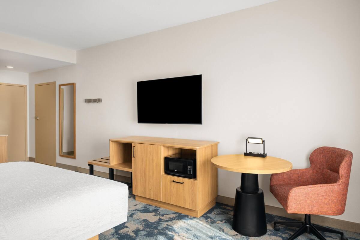 A hotel room with a bed, a wooden cabinet with a microwave, a television on the wall, and a round table with a chair.